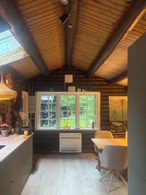 A cabin interior with a wooden ceiling, a kitchen area with a window, and a dining space with a table and chairs. - Timber-framed house with sedum roof in nature (Frederiksværk)