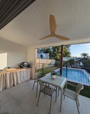 Ein überdachter Außenbereich mit Deckenventilator, Grill, Tisch mit Stühlen und Blick auf einen Swimmingpool.