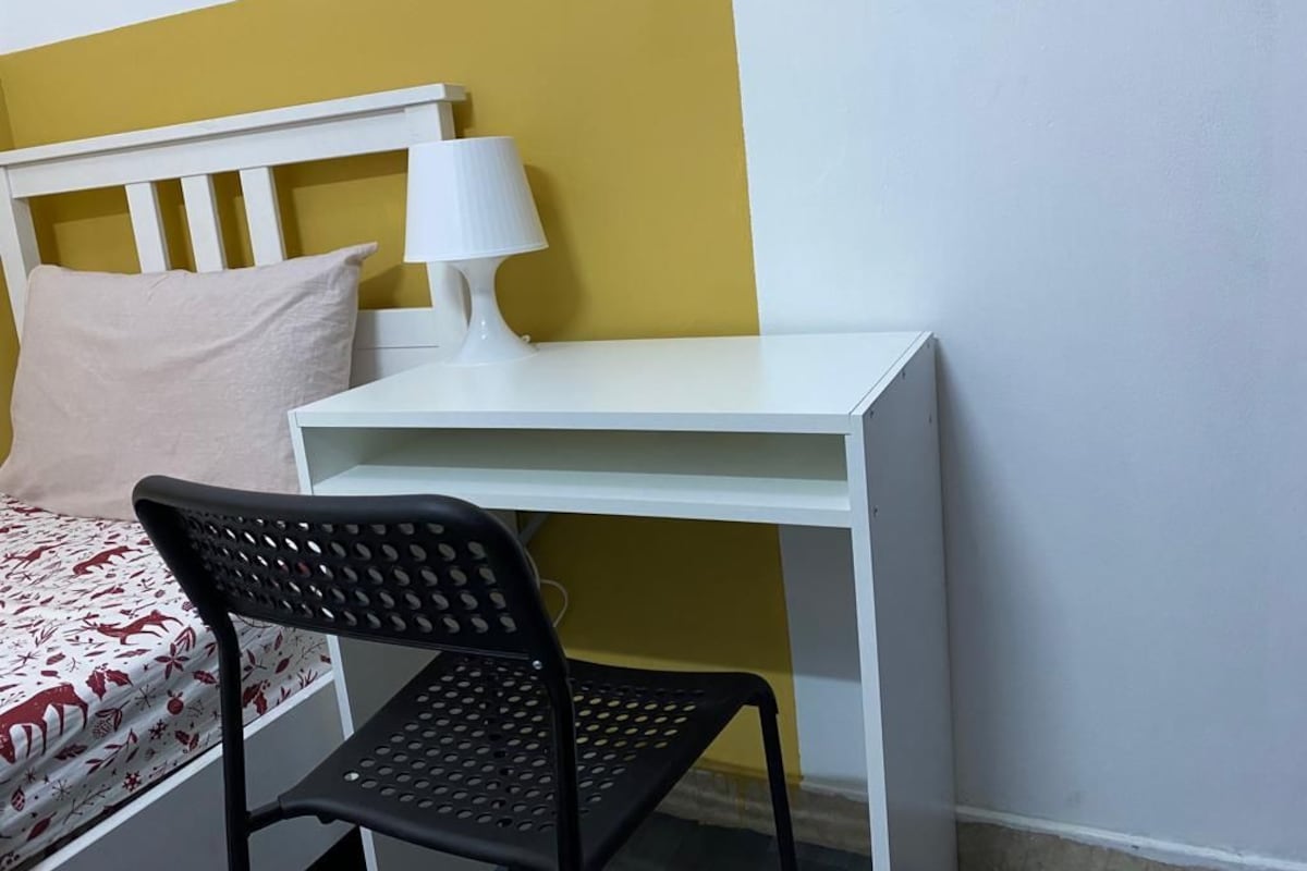 Een kamer met een bed, een wit bureau, een zwarte stoel en een lamp.