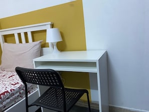 Een kamer met een bed, een wit bureau, een zwarte stoel en een lamp.