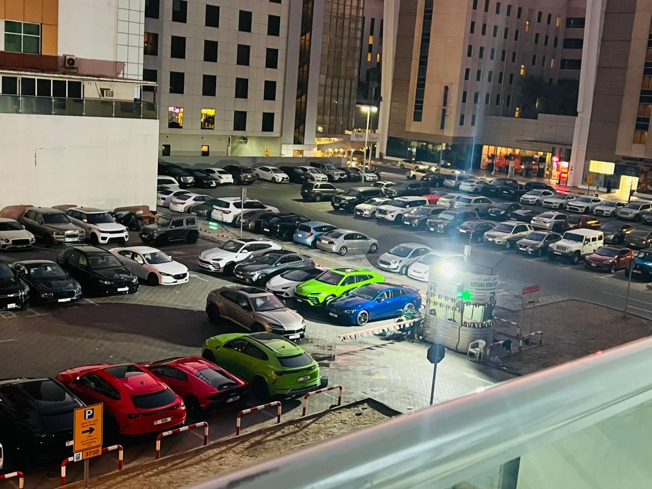 Een parkeerplaats met meerdere auto's, waaronder sportauto's, en een gebouw met verlichte ramen op de achtergrond.