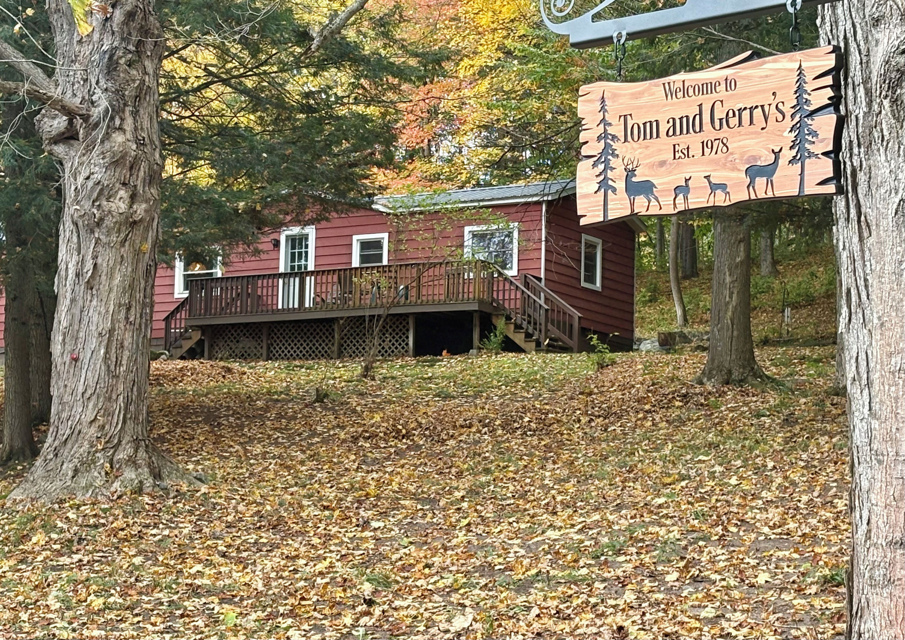 A wooden sign with a welcome message, a red building with a porch, and a forested area with fallen leaves.