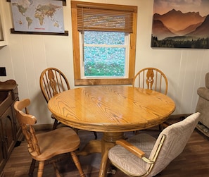 A dining area with a round wooden table, four chairs, a window with a view of greenery, a world map, and a mountain landscape painting. - 3-bedroom, 2 Bath with Infra-red Sauna mobile home, nestled among the trees. (Russell)
