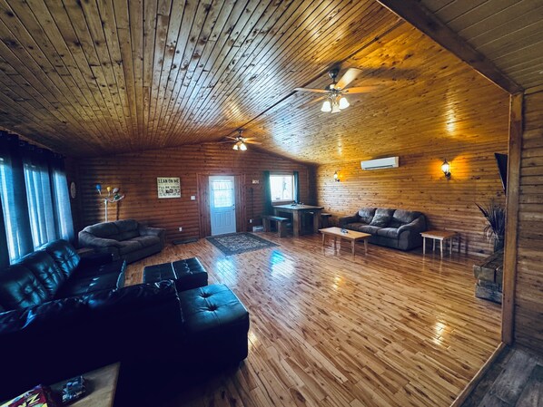 A log cabin interior with a wooden ceiling, a leather sofa, a dining table, and a ceiling fan.
