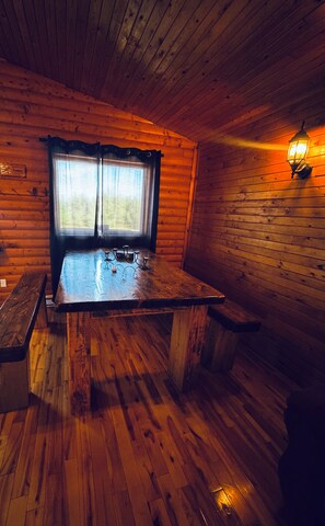 A cabin interior with a wooden table, benches, a window with curtains, and a wall-mounted light fixture.