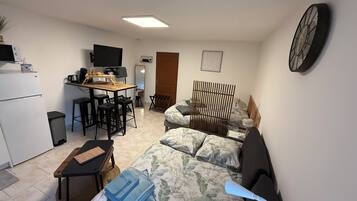A compact living space with a kitchenette, a bed with floral bedding, a desk with a chair, and a television.