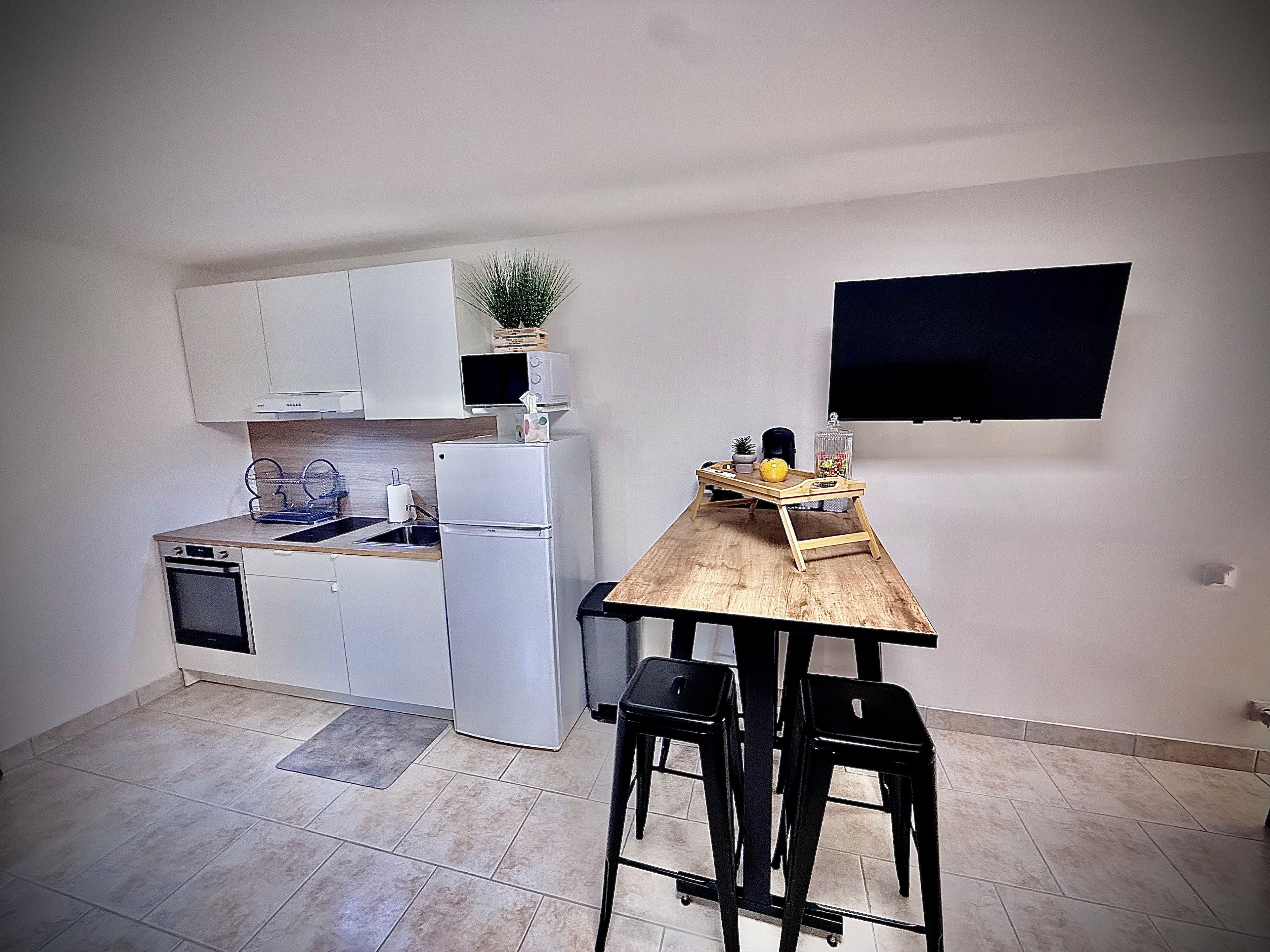 Een compacte keuken met witte kastjes, een ingebouwde magnetron en een kleine koelkast. Er is een houten tafel met twee zwarte stoelen en een dienblad met spullen erop.