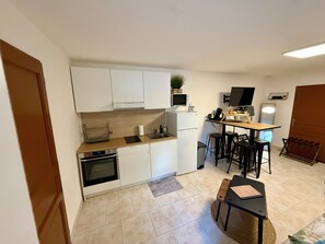 Een compacte keuken met witte kasten, een ingebouwde oven, een magnetron en een klein eethoekje met krukjes.