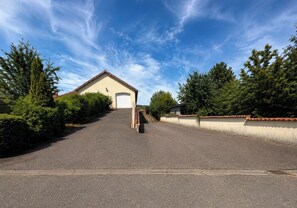 Een verharde oprit leidt naar een gebouw met een garagepoort, omgeven door groen en een heldere lucht.