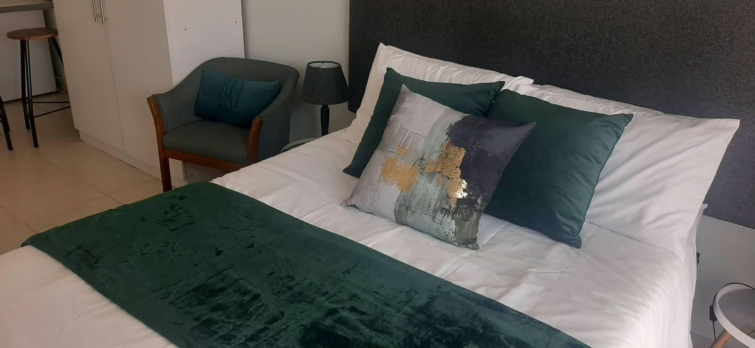 A bed with a green throw blanket, a pillow with a metallic design, and a chair with a dark cushion.