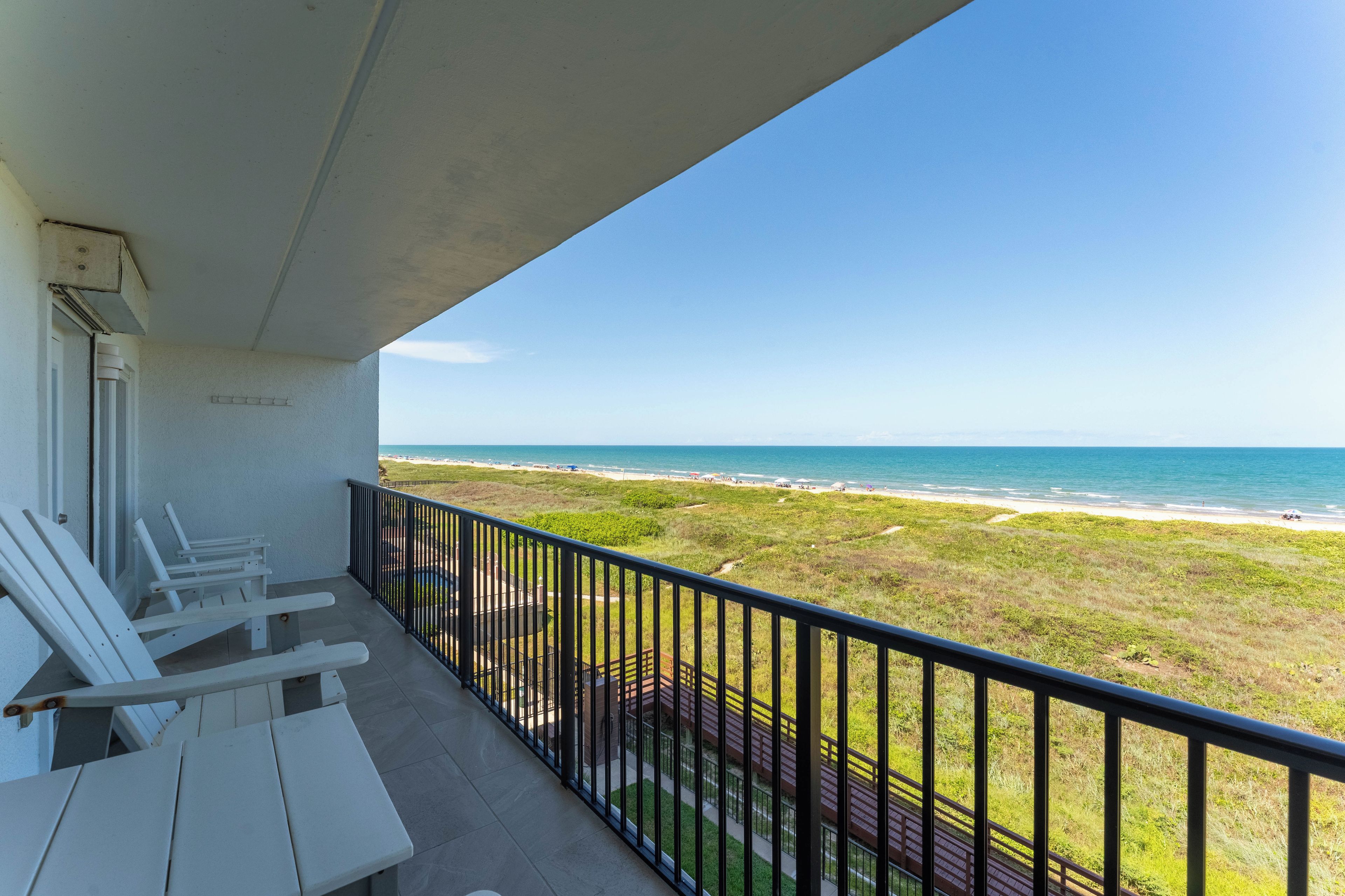 A balcony with white chairs and a view of the beach and ocean.