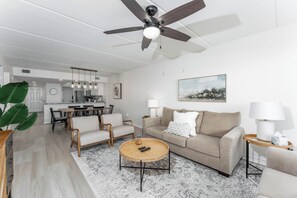 A modern living room with a sofa, coffee table, and chairs. A dining area with a table and chairs is visible in the background. - Seville 405 - Beachfront Condo Private Balcony, 2 Pools (South Padre Island)