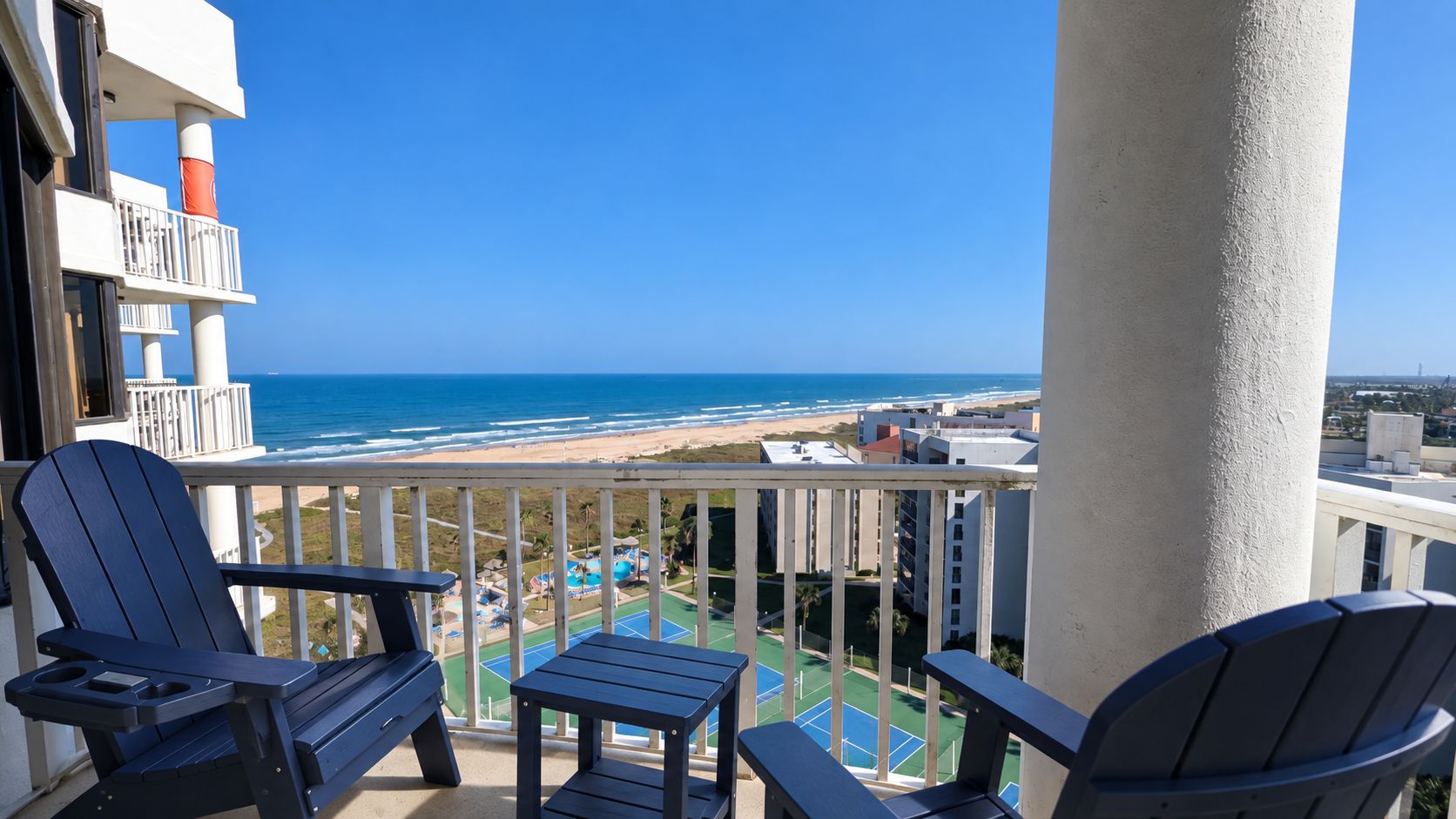 A balcony with two Adirondack chairs, a small table, and a view of the beach and ocean.