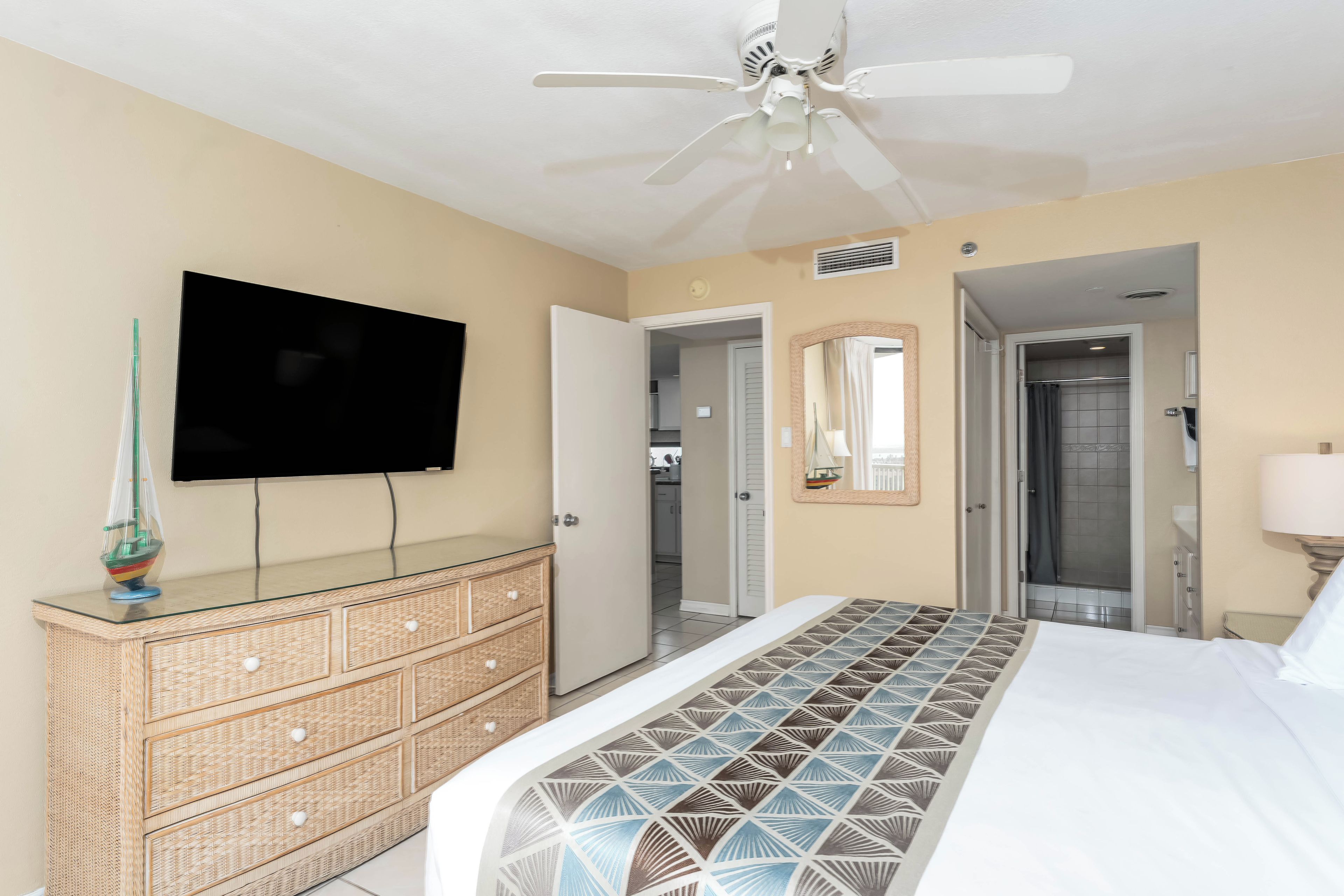 A bedroom with a bed, a television mounted on the wall, a ceiling fan, and a dresser.
