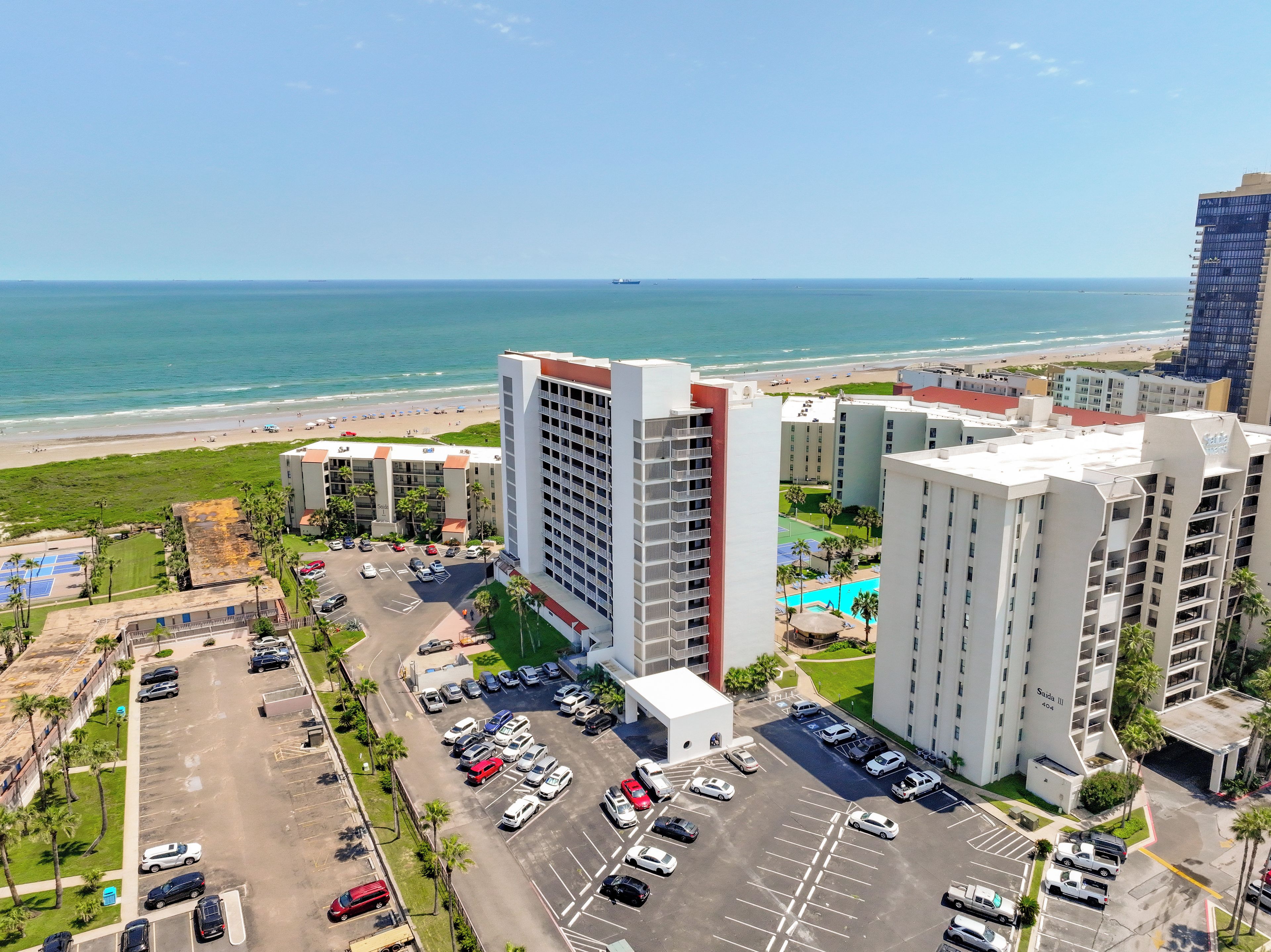A high-rise hotel with multiple floors, a parking lot, and a view of the beach.