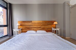 Ein Hotelzimmer mit einem Holzkopfteil, einem großen Bett mit weißer Bettwäsche, zwei Nachttischen mit Lampen und Blick durch ein Fenster.