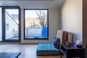 Ein Zimmer mit einem großen Fenster, einer blauen gepolsterten Bank, einem kleinen Tisch mit einer Lampe und einem Wandregal mit Dekorationselementen.