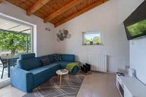 Ein modernes Wohnzimmer mit einem blauen Sofa, einem kleinen runden Tisch, einem an der Wand montierten Fernseher und einem Fenster mit Blick auf Grünflächen.