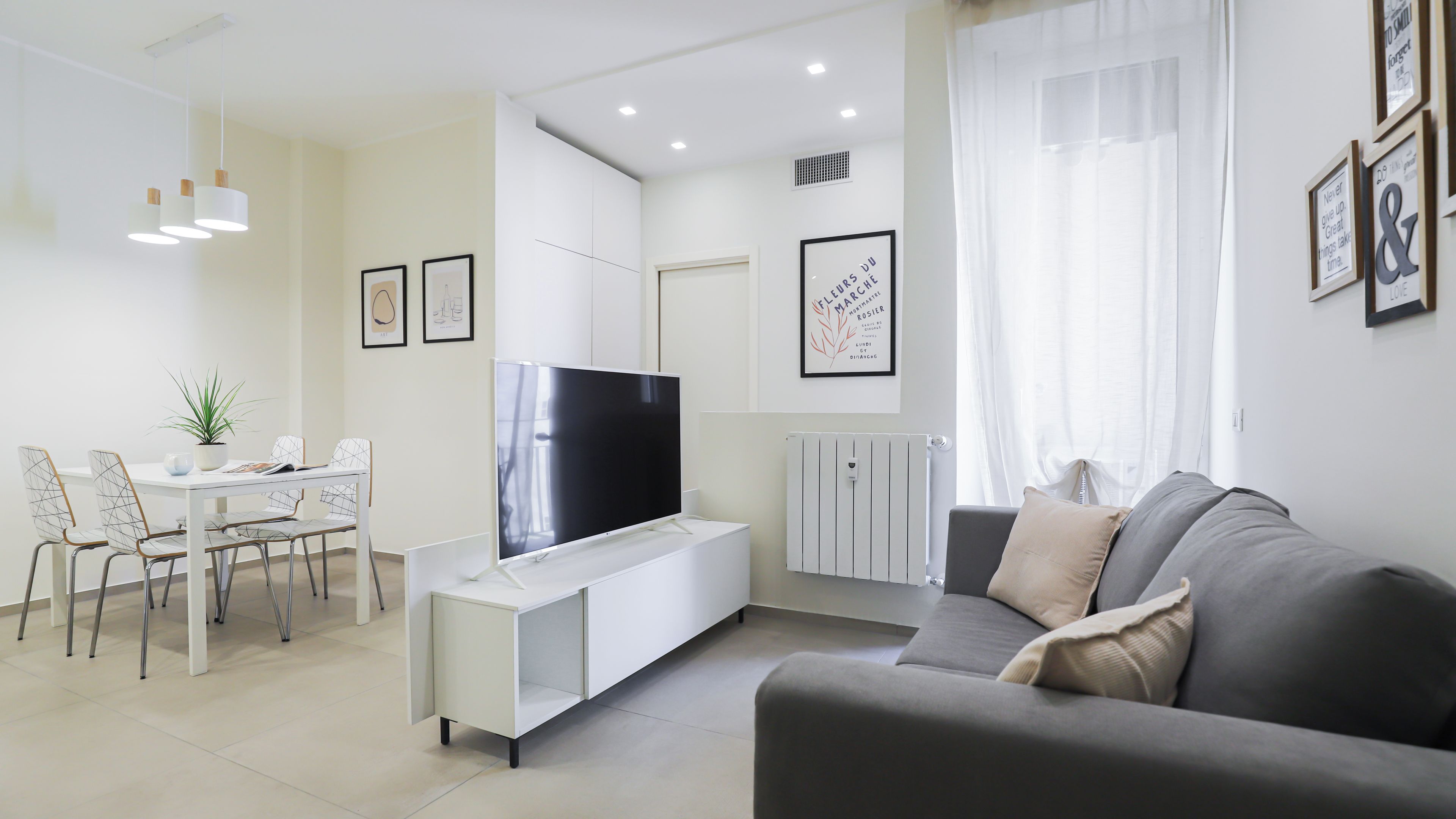 A modern living room with a flat-screen TV, a grey sofa, a dining table with chairs, and a potted plant.
