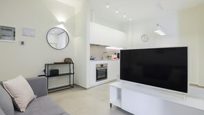 A modern living room with a flat-screen TV, a grey sofa, a coffee table, a kitchen area with white cabinets, and a round mirror. - Italianway Comfort - Soperga 15 (Milan)