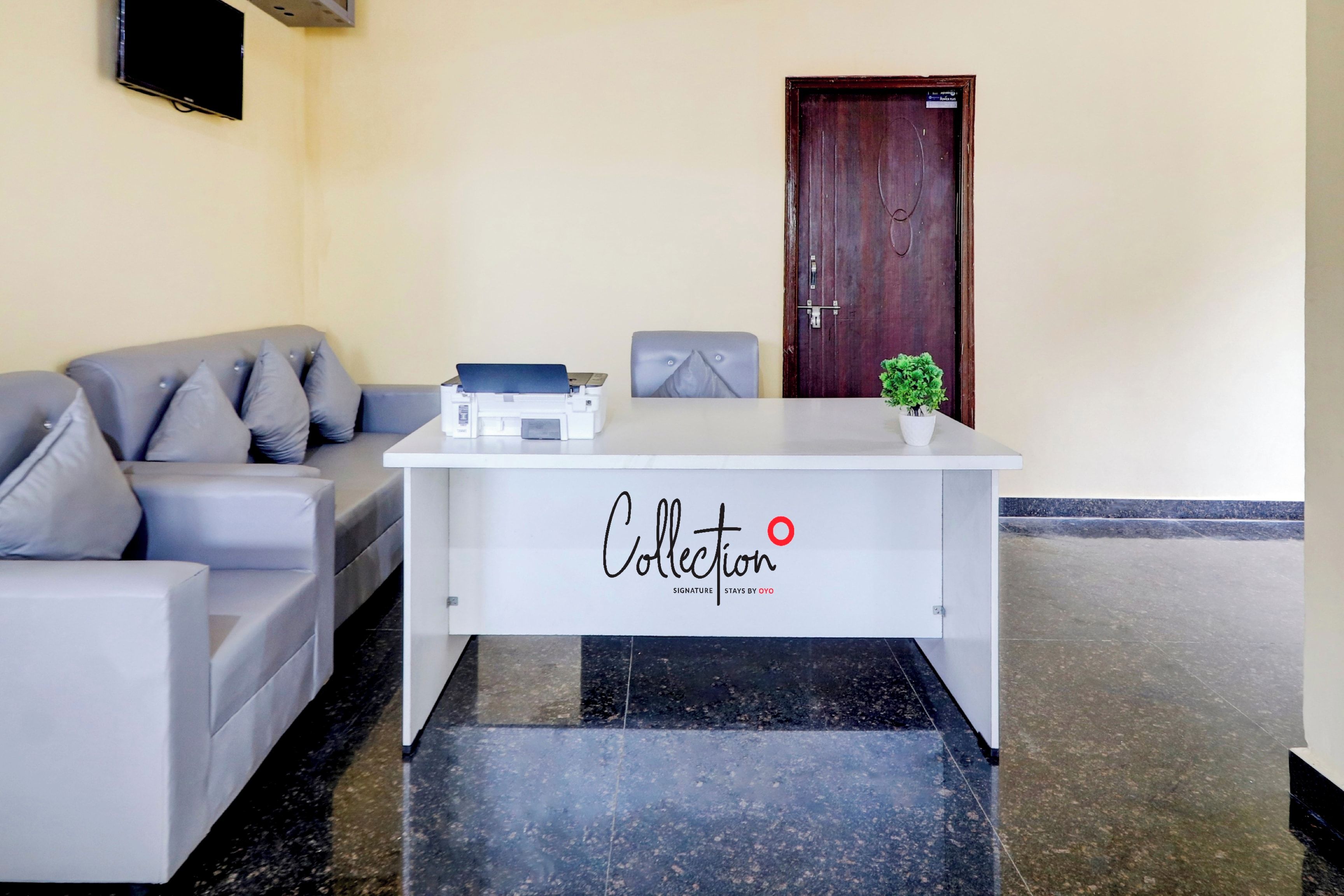 Una recepción con un cartel que dice 'Collection Signature Stays by OYO' en el vestíbulo de un hotel, con un sofá gris y una puerta de madera.