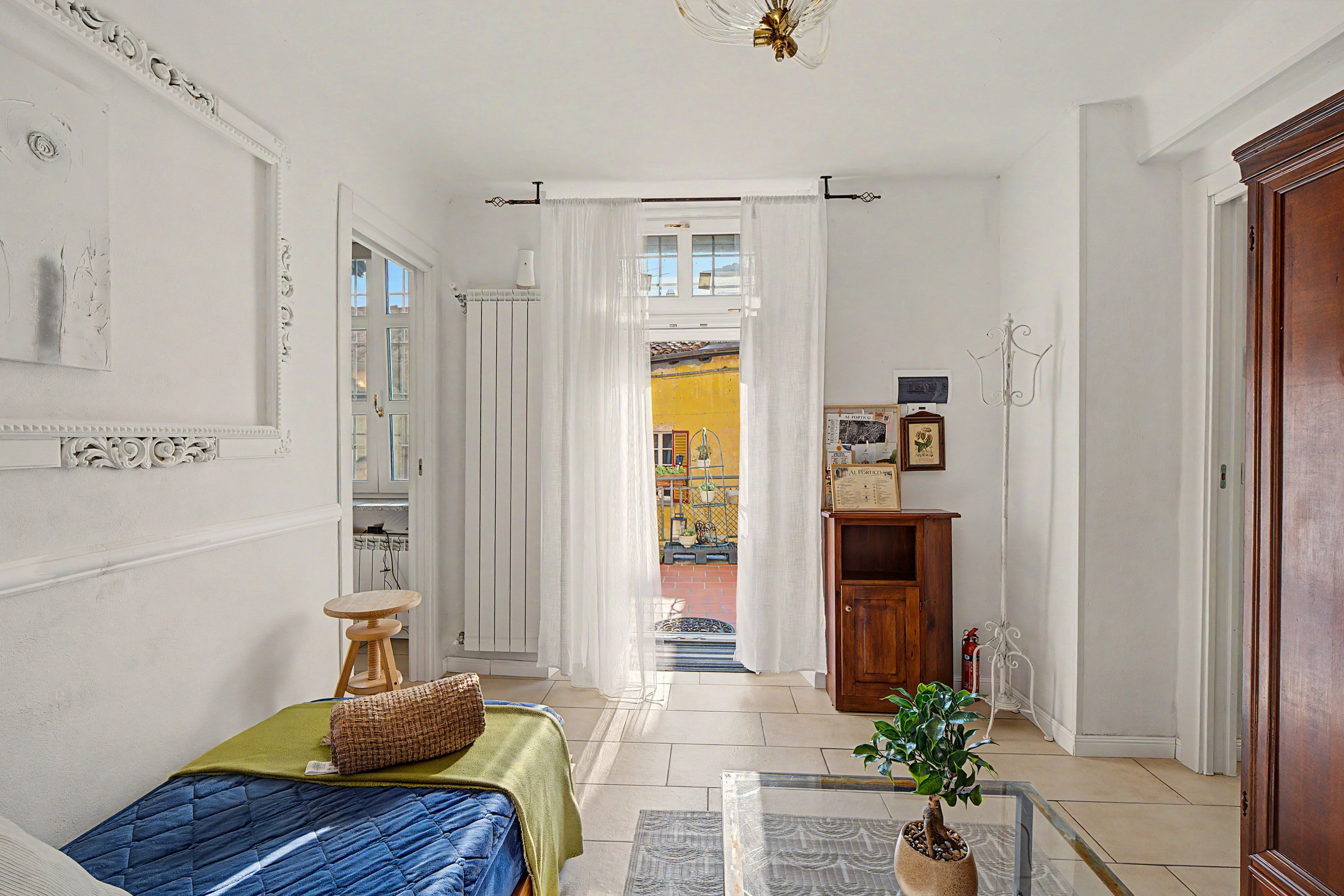 Una habitación con cama, una mesita, una planta en maceta y vistas a un espacio exterior por un vano de puerta.