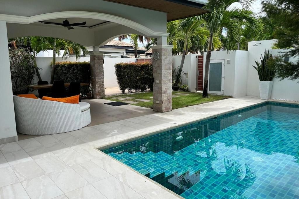 Una piscina con solárium azulejado, una zona de estar techada con cojines y una palmera de fondo.