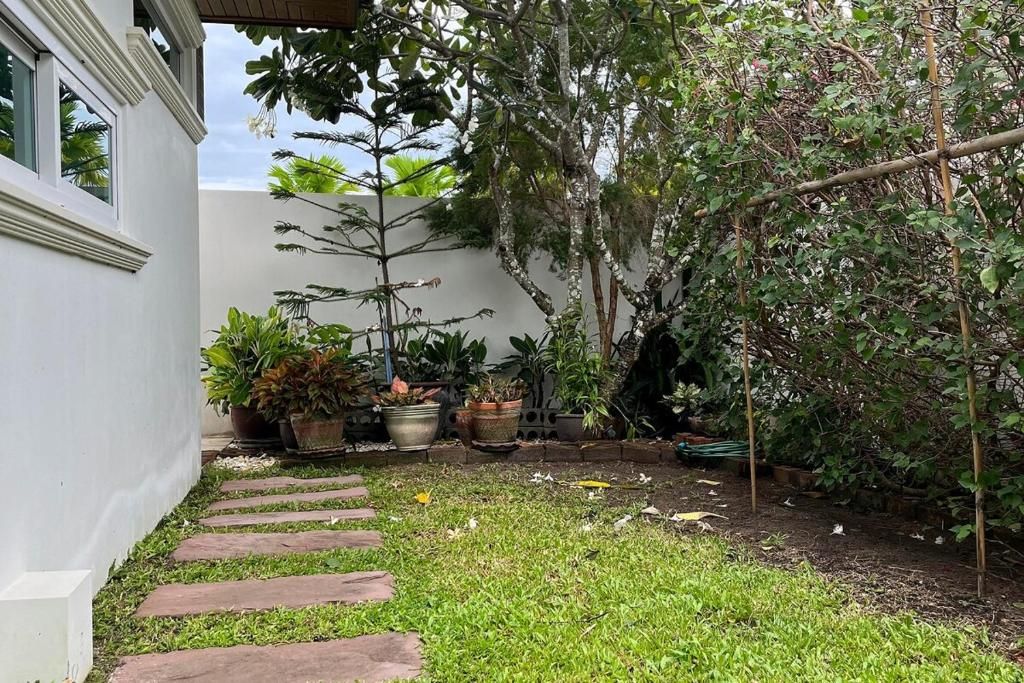 Un jardin avec un chemin de pierre menant à un bâtiment blanc, entouré de différentes plantes en pot et d’arbres.