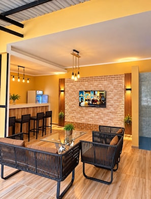 A modern living area with a glass-top coffee table, wicker furniture, a flat-screen TV mounted on a brick wall, and a kitchen area with bar stools.