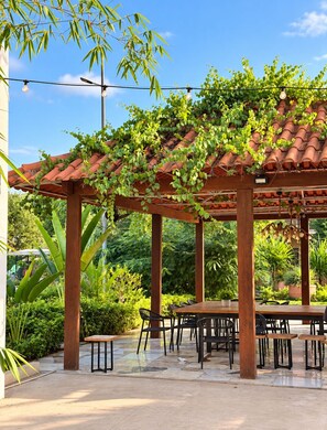Een terras buiten met een houten pergola, een betegelde vloer en een tafel met stoelen.