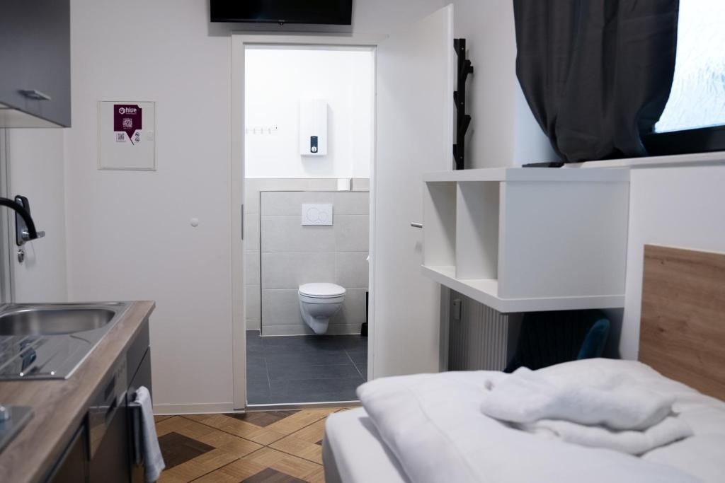 A hotel room with a bed, a small kitchenette, a bathroom with a toilet and sink, and a TV mounted on the wall.