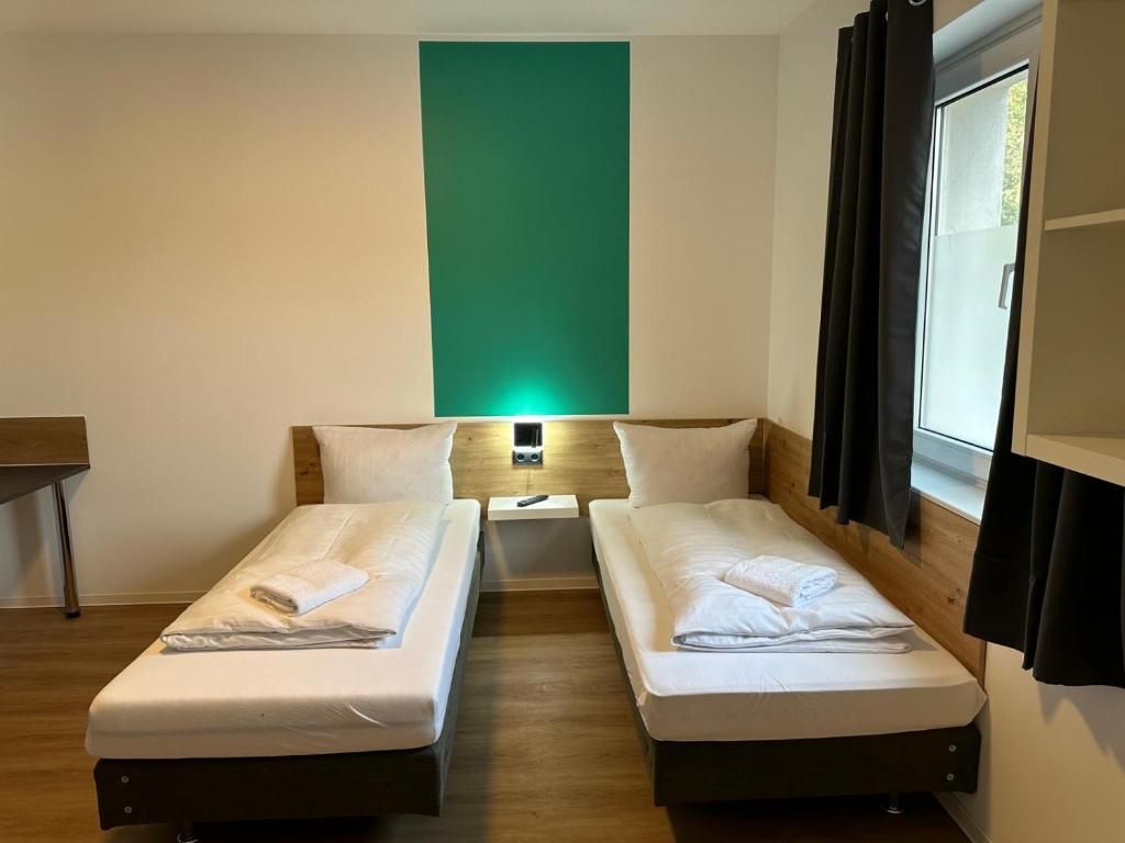 A hotel room with two single beds, a small bedside table with a lamp, a green accent wall, and a window with curtains.