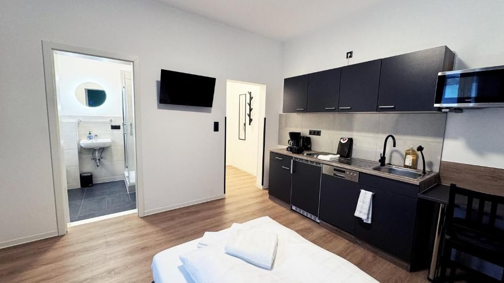 A compact hotel room with a kitchenette, a bed with a white towel, a television mounted above the counter, and a door leading to a bathroom.