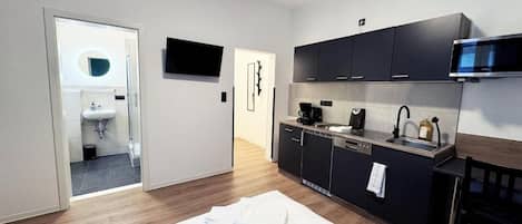 A compact hotel room with a kitchenette, a bed with a white towel, a television mounted above the counter, and a door leading to a bathroom.