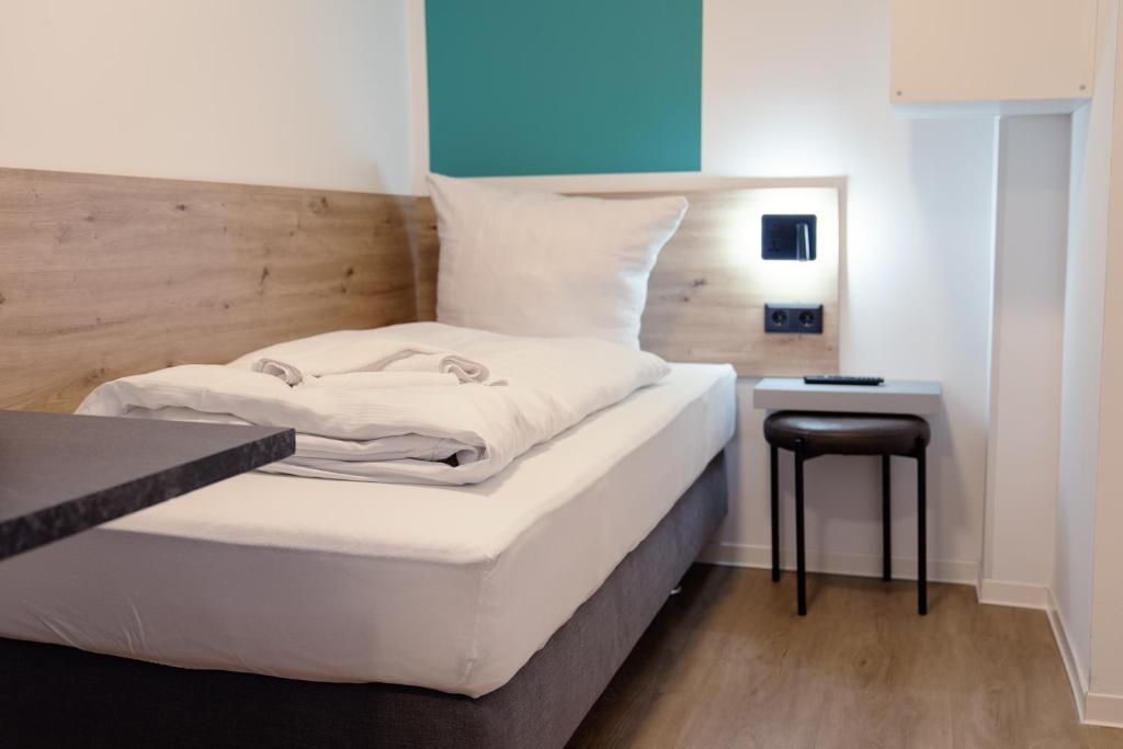 Ein ordentlich bezogenes Bett mit weißer Decke und Kissen, ein kleiner Nachttisch mit Lampe sowie ein schwarzer Stuhl.