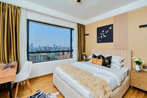 Een moderne slaapkamer met een groot raam, een bed met decoratieve kussens, een bureau met een laptop en uitzicht op de stad.