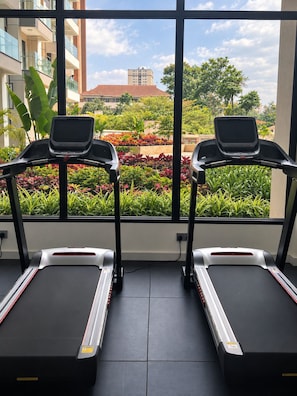 Ein Fitnessraum mit zwei Laufbändern, einem Fenster und Blick auf ein Gebäude und Grünflächen.