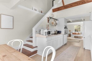 A modern kitchen with white cabinetry, a wooden dining table, and a staircase leading to a loft area. - 2BR Coastal Retreat w/ River & Sea Views (Jenner)