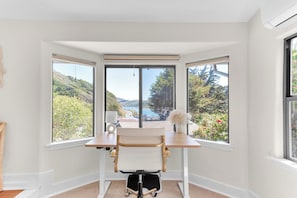 Une pièce d’angle équipée d’un bureau, d’une chaise et de grandes fenêtres donnant sur une colline et un plan d’eau.