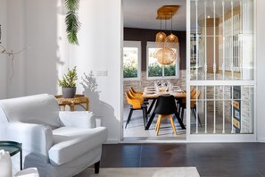 Een moderne woonkamer met een witte bank, een eethoek met een houten tafel en stoelen, en uitzicht op de omgeving door grote ramen.