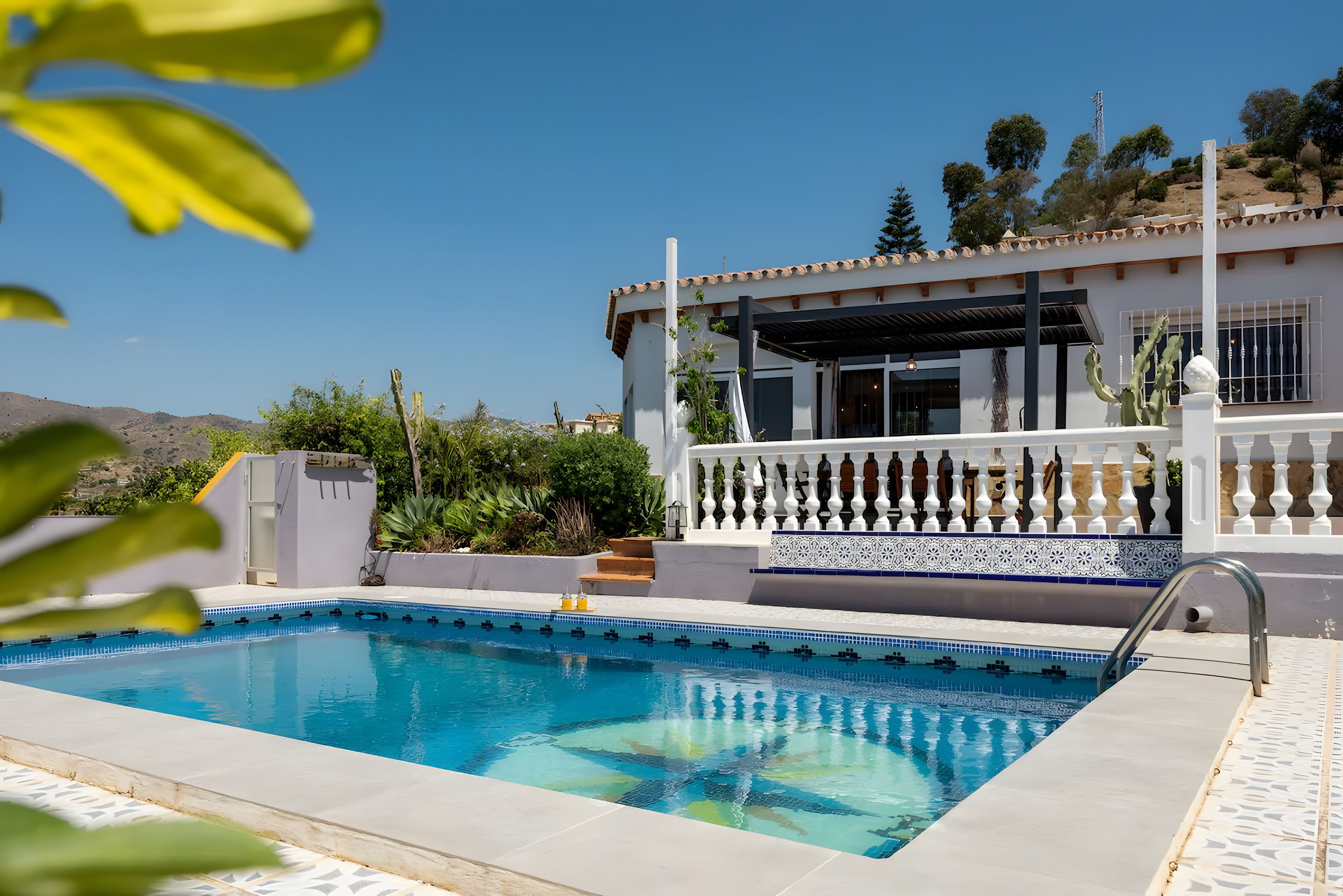 En swimmingpool med en klar blå vandoverflade, omgivet af en hvid bygning med balkon og udsigt til bakker i baggrunden.