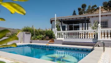 En swimmingpool med en klar blå vandoverflade, omgivet af en hvid bygning med balkon og udsigt til bakker i baggrunden.