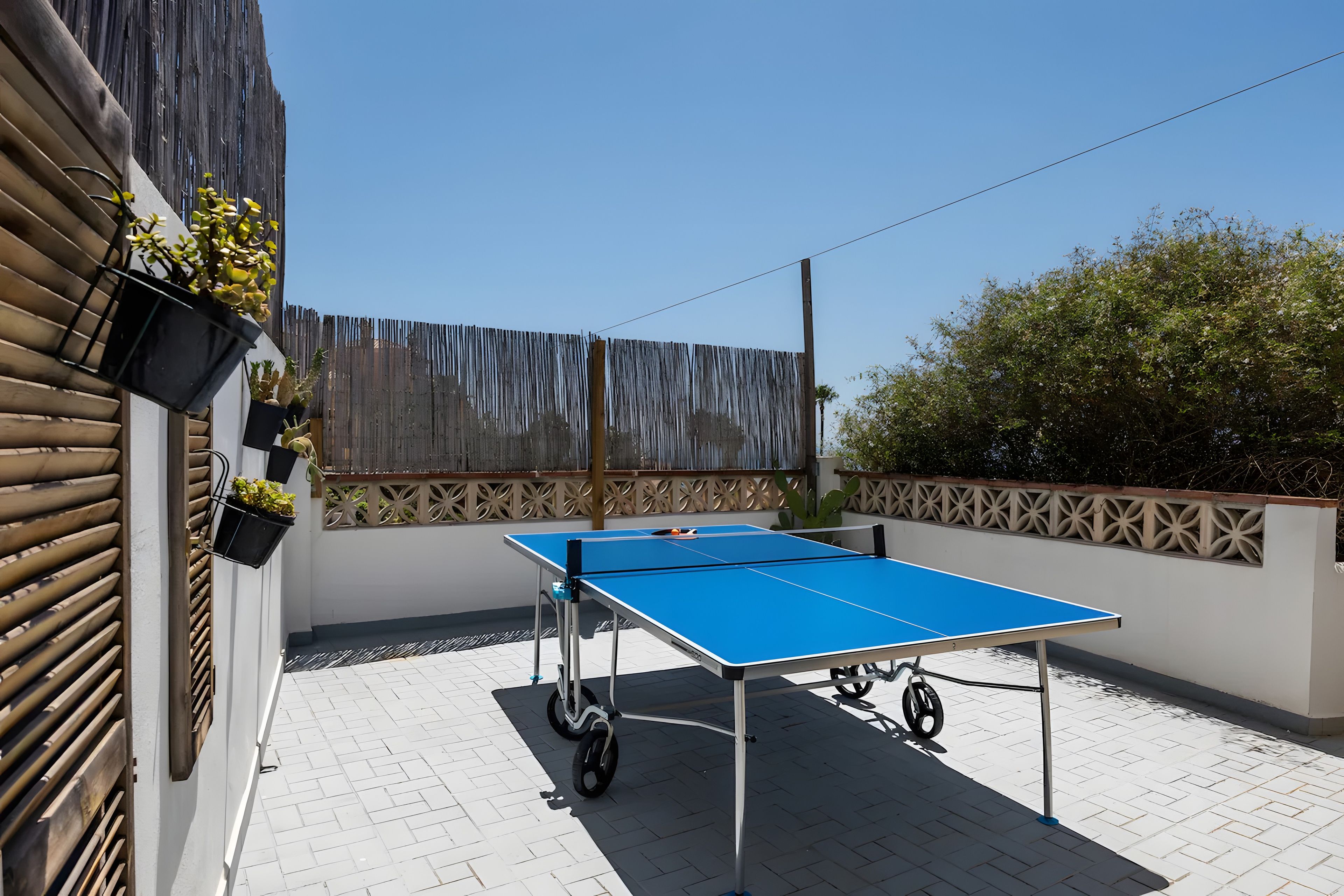 Mesa de ping pong al aire libre en un patio empedrado con cercado de madera y plantas en macetas.