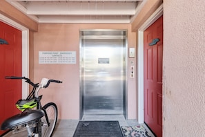 An elevator with emergency contacts listed on the wall, a bicycle parked to the left, and a red door on the right. - Oceanfront Views, Beach Steps Away, Walk Everywhere by Tybee Cottages (Tybee Island)