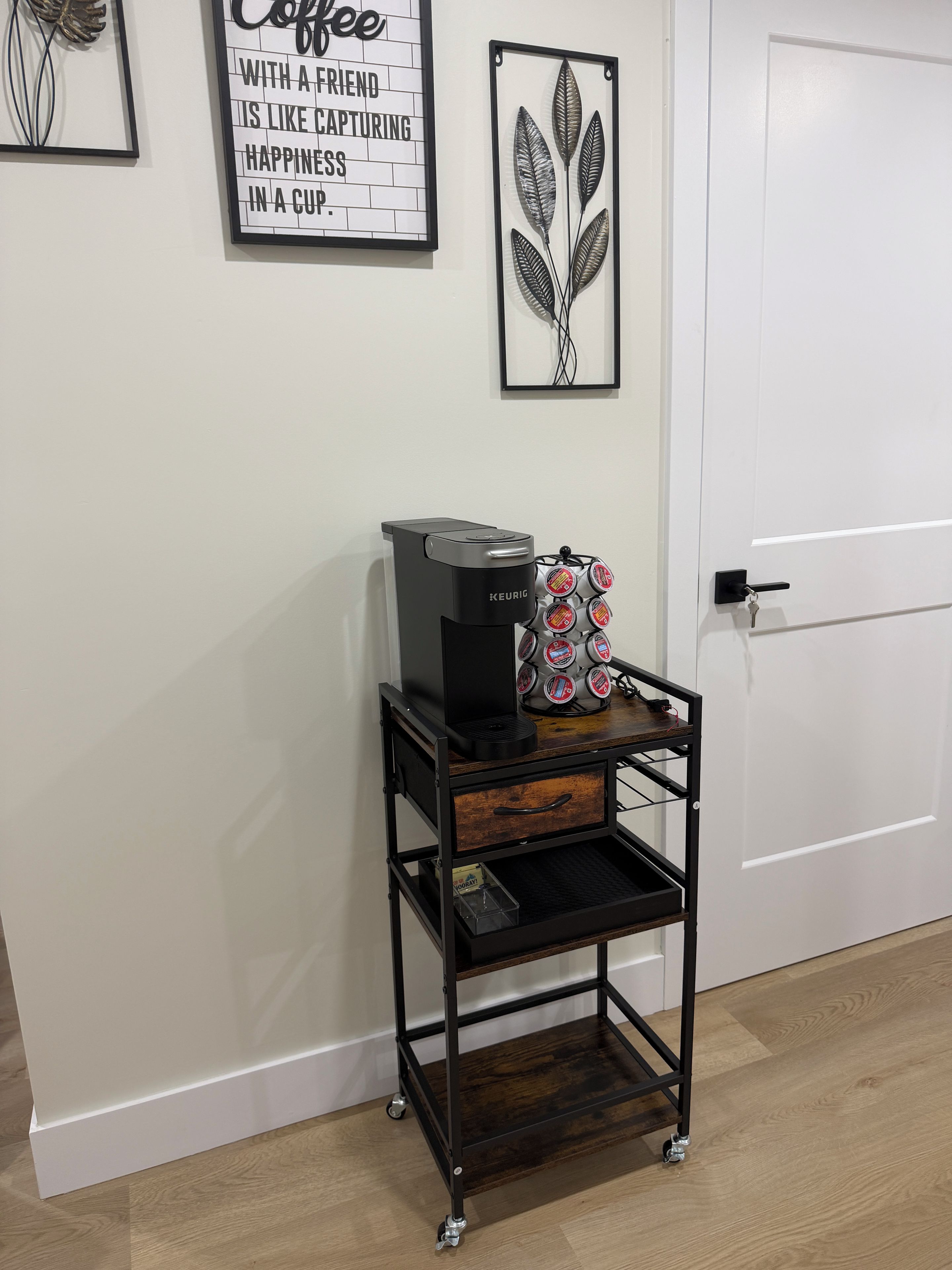 Un espace dédié au café, comprenant une machine Keurig, un porte-tasse, un tiroir en bois et une étagère en métal, le tout installé sur un chariot en métal noir.