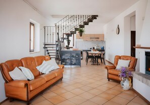 A modern living room with a brown leather sofa, a dining area with a wooden table, and a kitchen with white cabinets and a black staircase. - Holiday Home 'Farmhouse with Garden and Pool' with Mountain View, Private Pool and Wi-Fi (Cingoli)