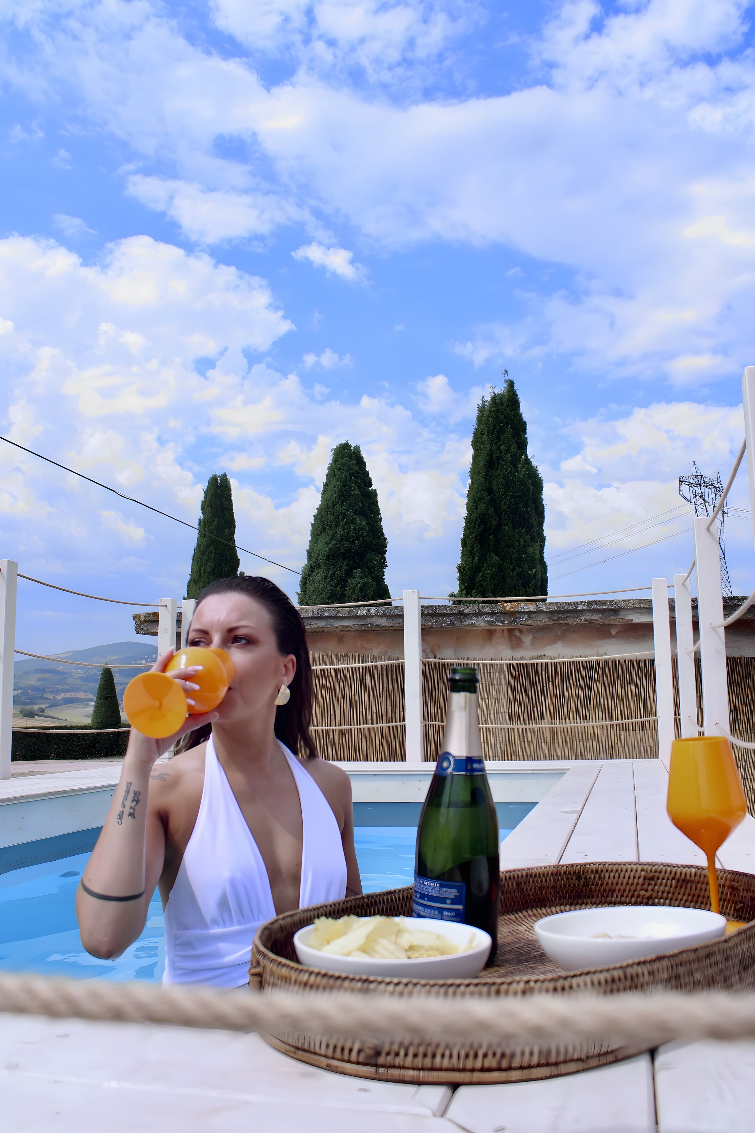 A person by a pool with a bottle of wine and a glass of orange juice.