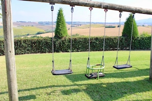 A wooden swing set with two swings in a grassy field with a hedge and distant hills. - Holiday Home 'Farmhouse with Garden and Pool' with Mountain View, Private Pool and Wi-Fi (Cingoli)