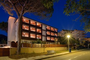 Un edificio de varios pisos con ventanas iluminadas de noche, una banca en la acera y un farol.