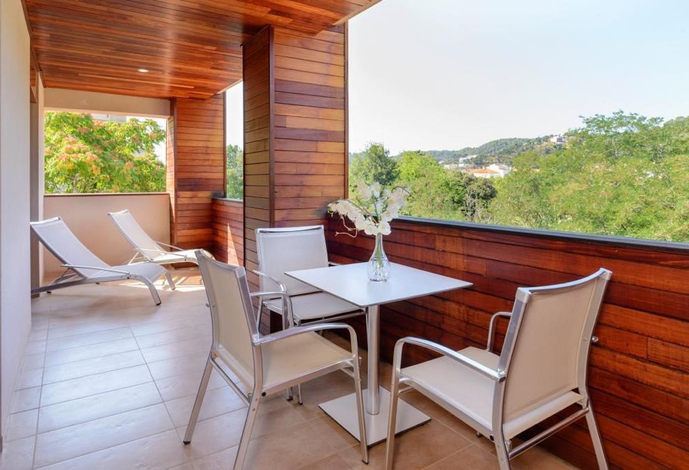 Ein Balkon mit Holzboden, weißen Stühlen, einem Tisch und einer Blumenvase.